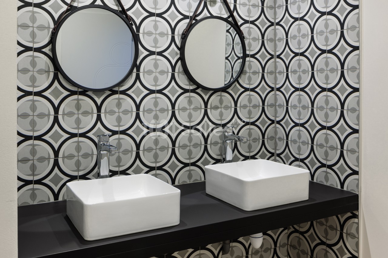 Deux lavabos blancs avec robinets modernes sur un plan noir, devant un mur carrelé à motifs géométriques.