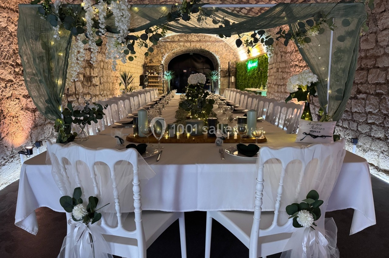 Table de banquet décorée avec des fleurs, bougies et guirlandes lumineuses, installée dans une salle voûtée en pierre.