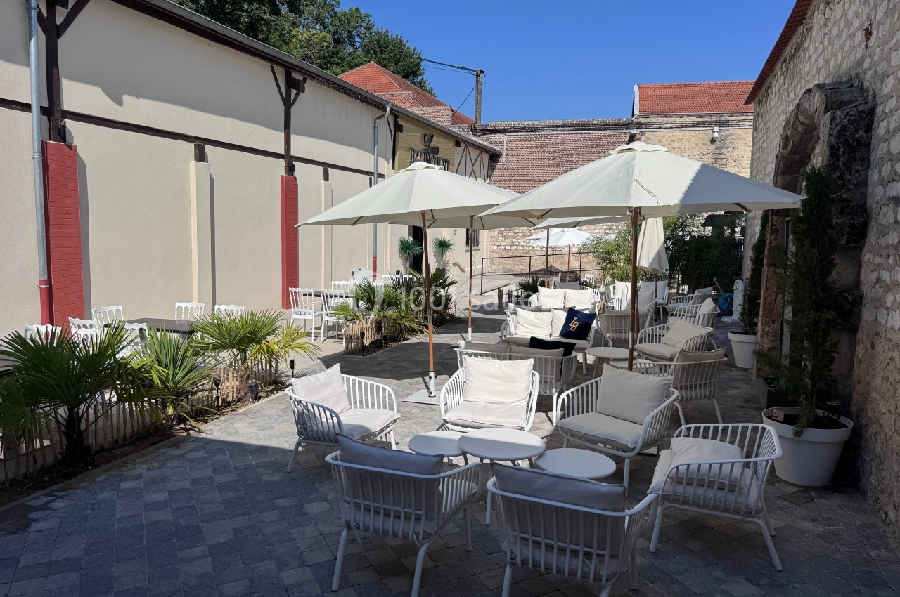 Terrasse extérieure avec tables, chaises blanches et parasols, entourée de murs en pierre et végétation.