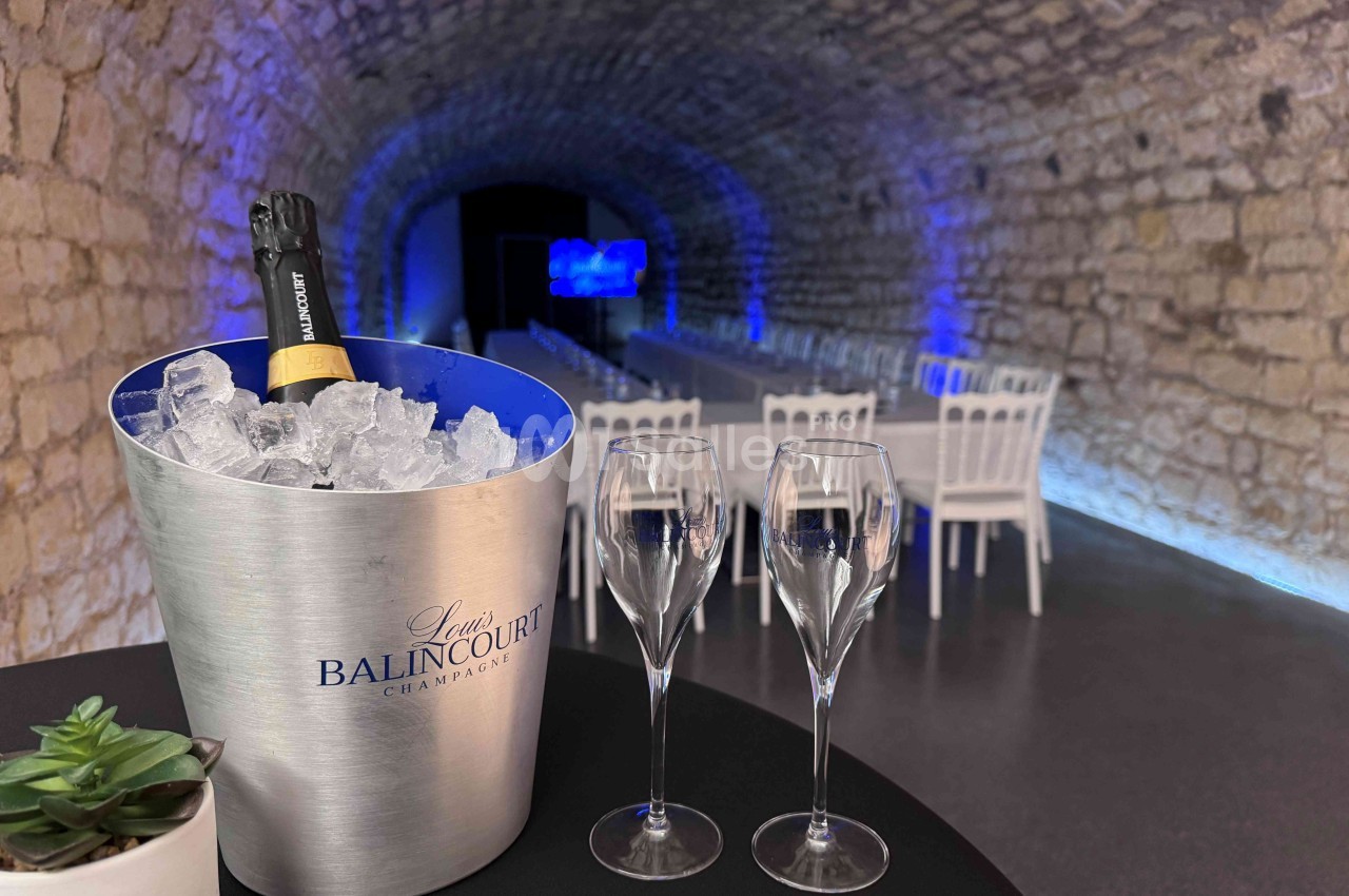 Seau à champagne avec bouteille sur glace, deux flûtes vides et une plante, dans une salle voûtée éclairée en bleu.
