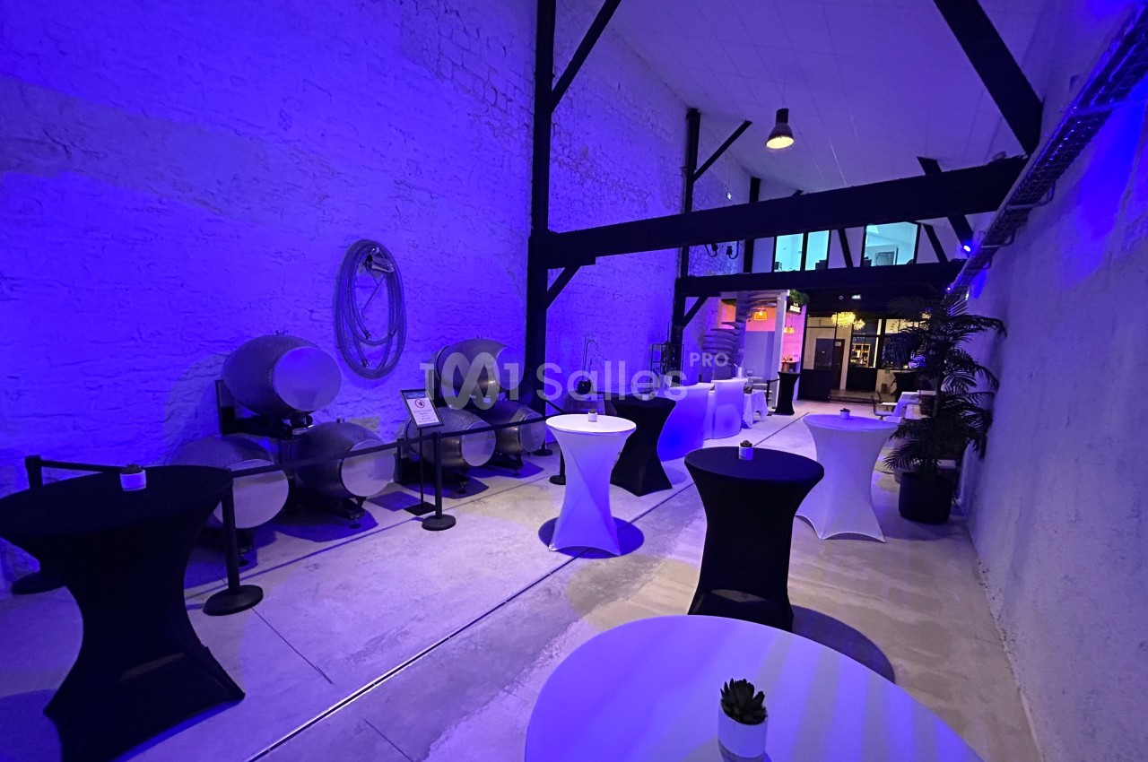 Salle avec murs en pierre, tonneaux en arrière-plan et tables hautes éclairées par une lumière bleue.
