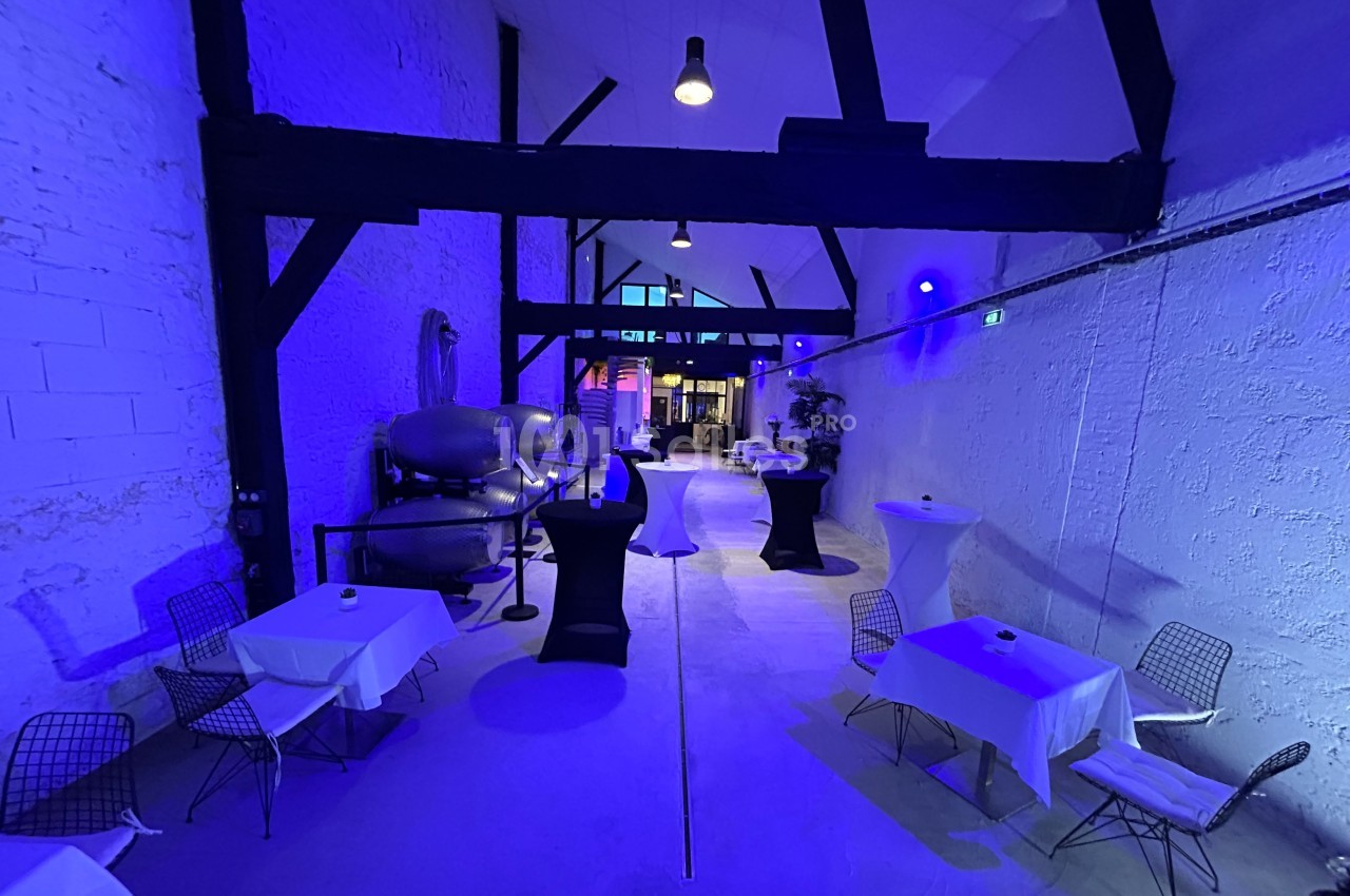 Salle industrielle avec murs blancs, éclairage bleu, tables dressées et chaises modernes, ambiance épurée et contemporaine.