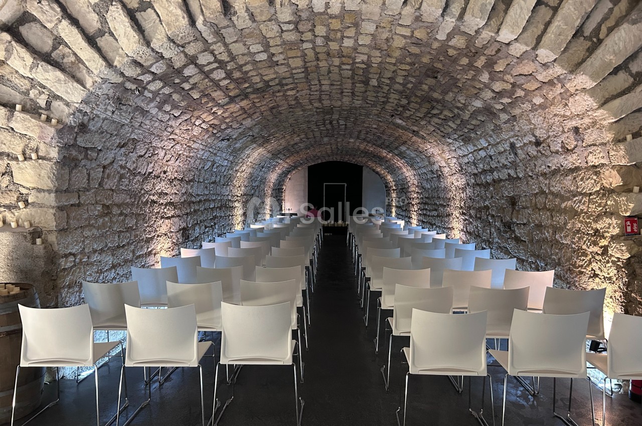Salle voûtée en pierre aménagée avec des rangées de chaises blanches alignées face à un espace de présentation.