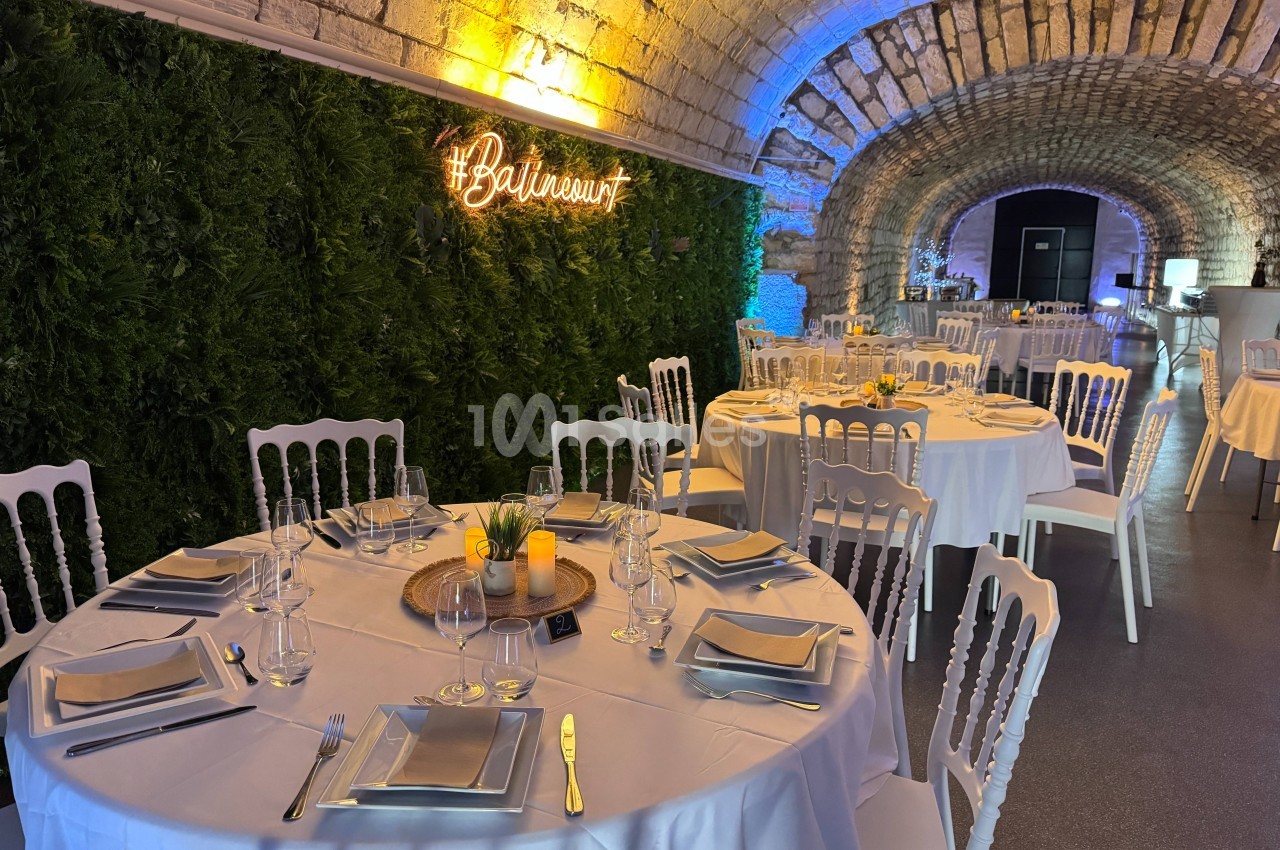 Salle voûtée décorée avec des tables rondes dressées, nappes blanches, mur végétal et enseigne lumineuse.