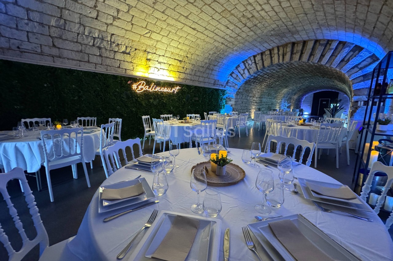 Salle voûtée en pierre décorée pour un événement, avec tables dressées et éclairage tamisé bleu et jaune.