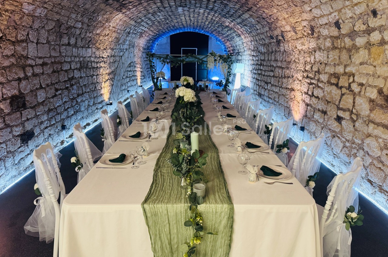 Salle voûtée en pierre aménagée avec une longue table décorée de nappes blanches, verdure et fleurs.