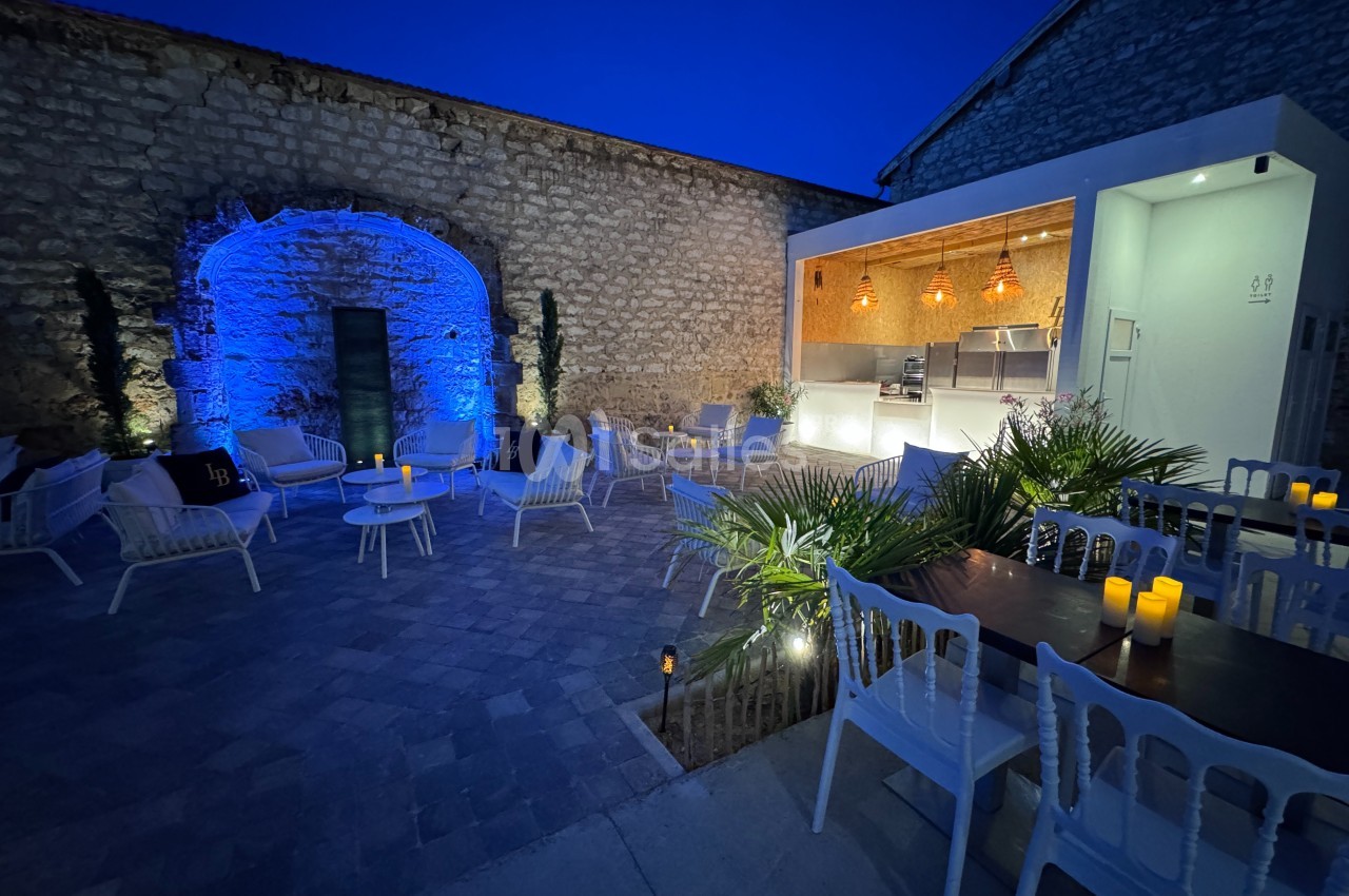 Cour extérieure en soirée avec éclairage bleu, mobilier de jardin, plantes et un espace cuisine illuminé.