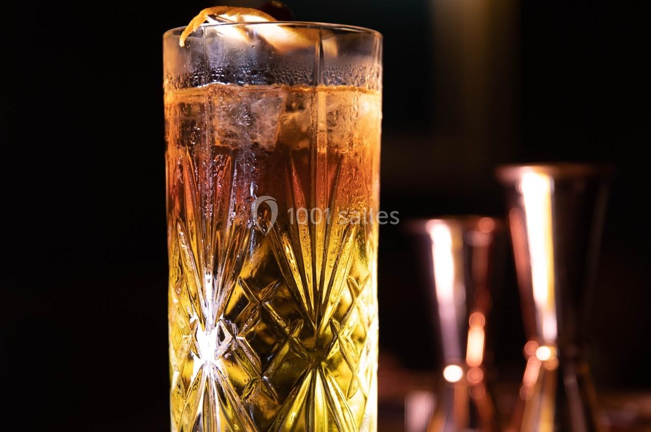 Verre à cocktail gravé rempli d'une boisson ambrée avec des glaçons et un zeste d'agrume, éclairé en ambiance tamisée.