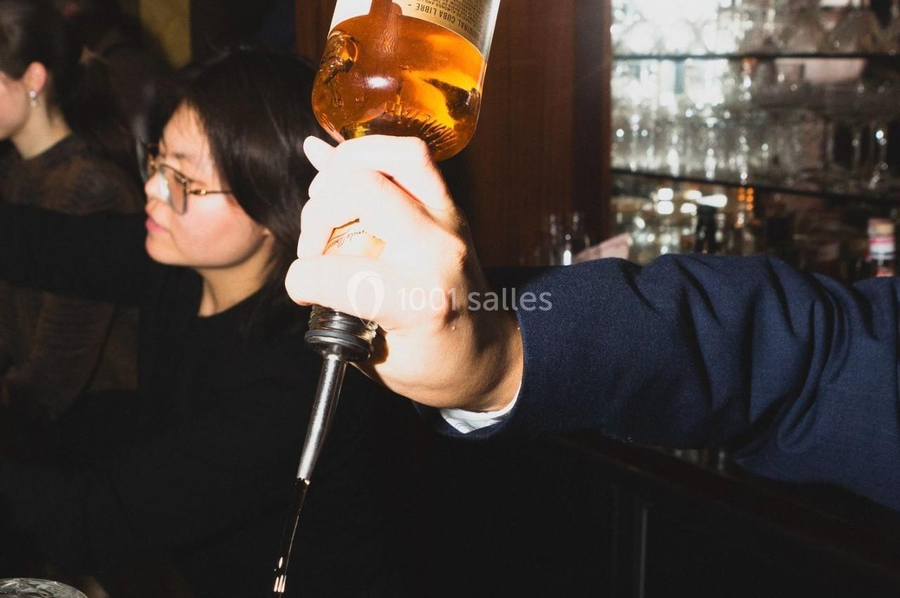 Une main verse de l'alcool d'une bouteille dans un verre, avec un bar en arrière-plan.