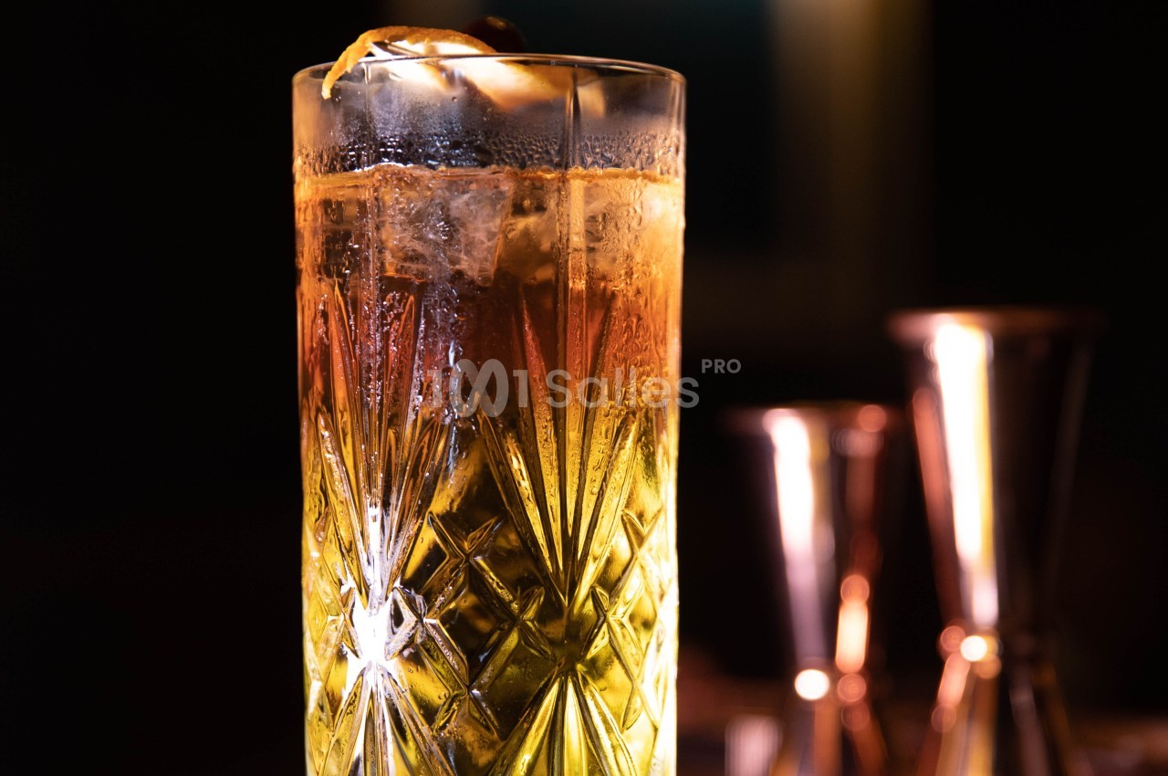 Verre à cocktail en cristal rempli d'une boisson ambrée avec glaçons, décoré d'un zeste d'agrume et d'une cerise.