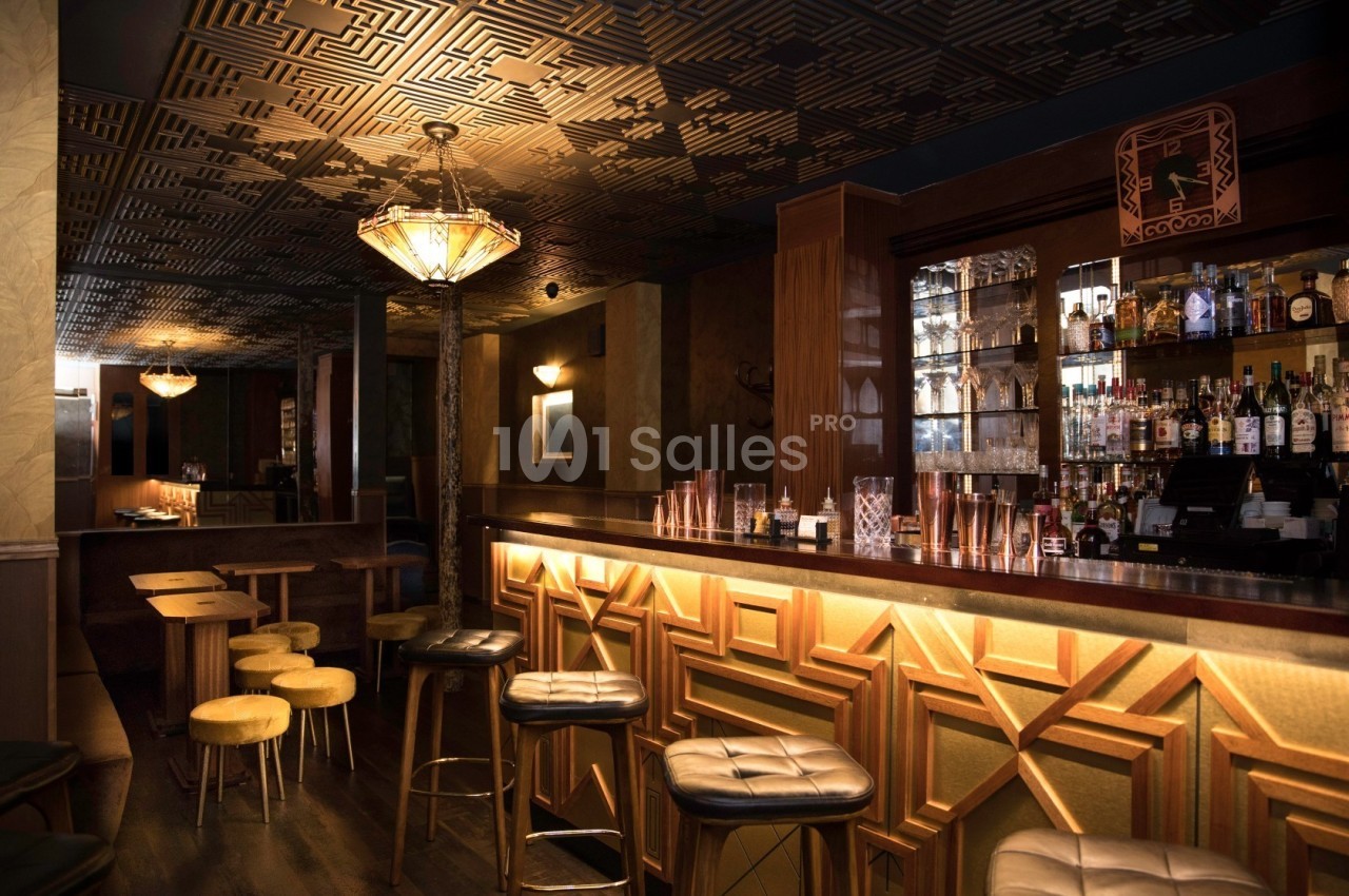Intérieur d'un bar avec un comptoir éclairé, des tabourets, des étagères de bouteilles et un plafond décoratif.