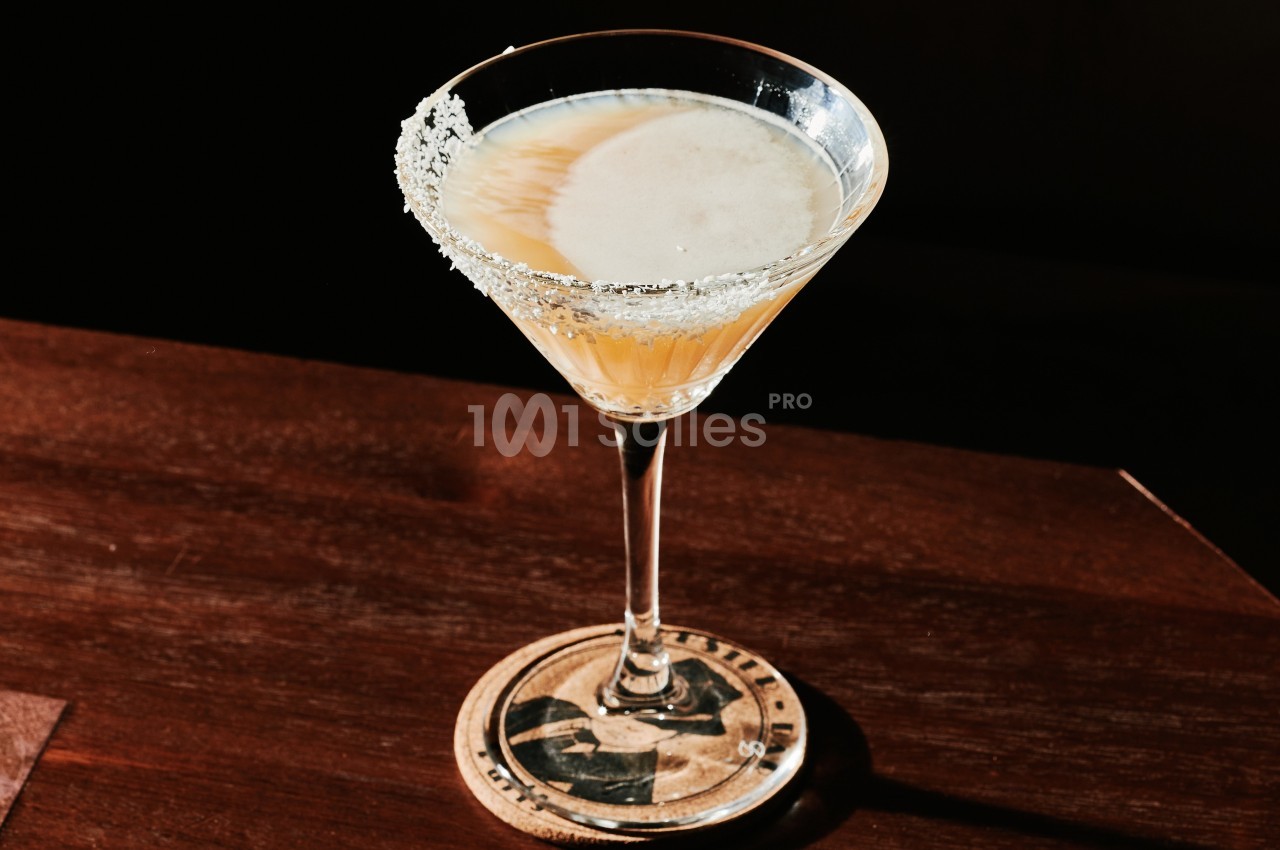 Verre à cocktail avec bord salé, posé sur un sous-verre en bois, éclairé par une lumière tamisée.