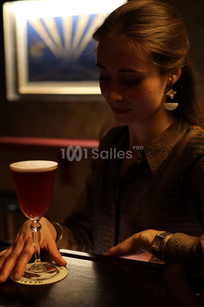 Une femme assise dans un bar sombre tient un menu près d'un cocktail rouge servi dans un verre élégant.