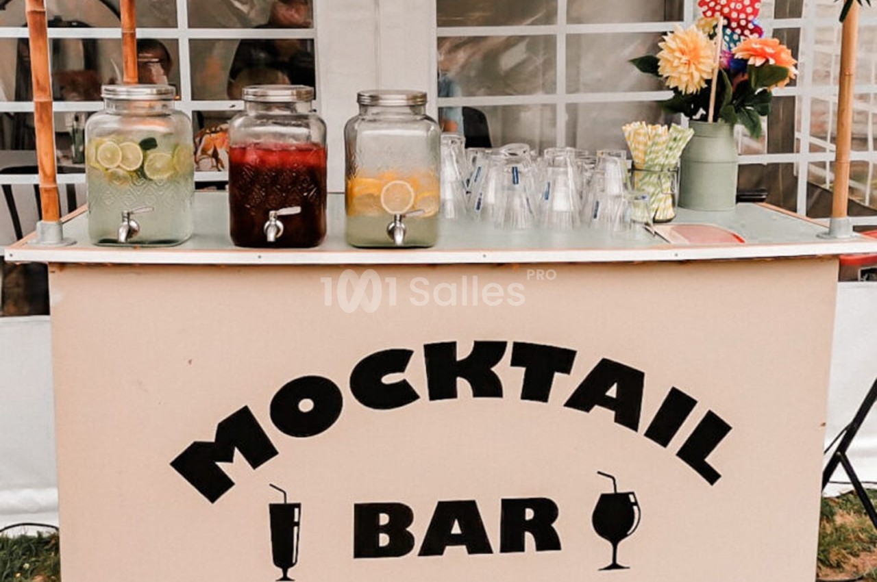 Bar à mocktails avec trois distributeurs de boissons colorées, verres en plastique et décorations florales en arrière-plan.
