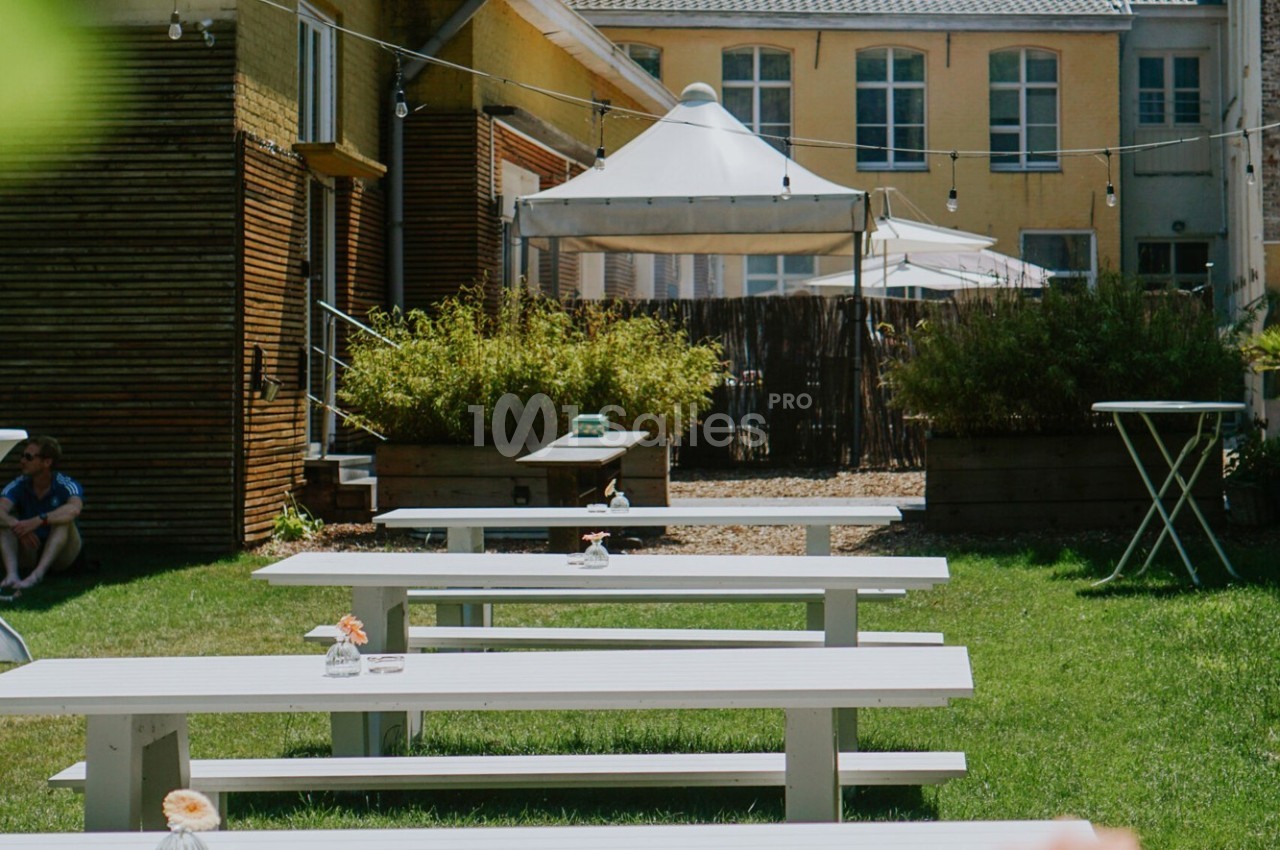 Tables de pique-nique blanches alignées sur une pelouse, entourées de bâtiments et d'un espace ombragé avec des parasols.
