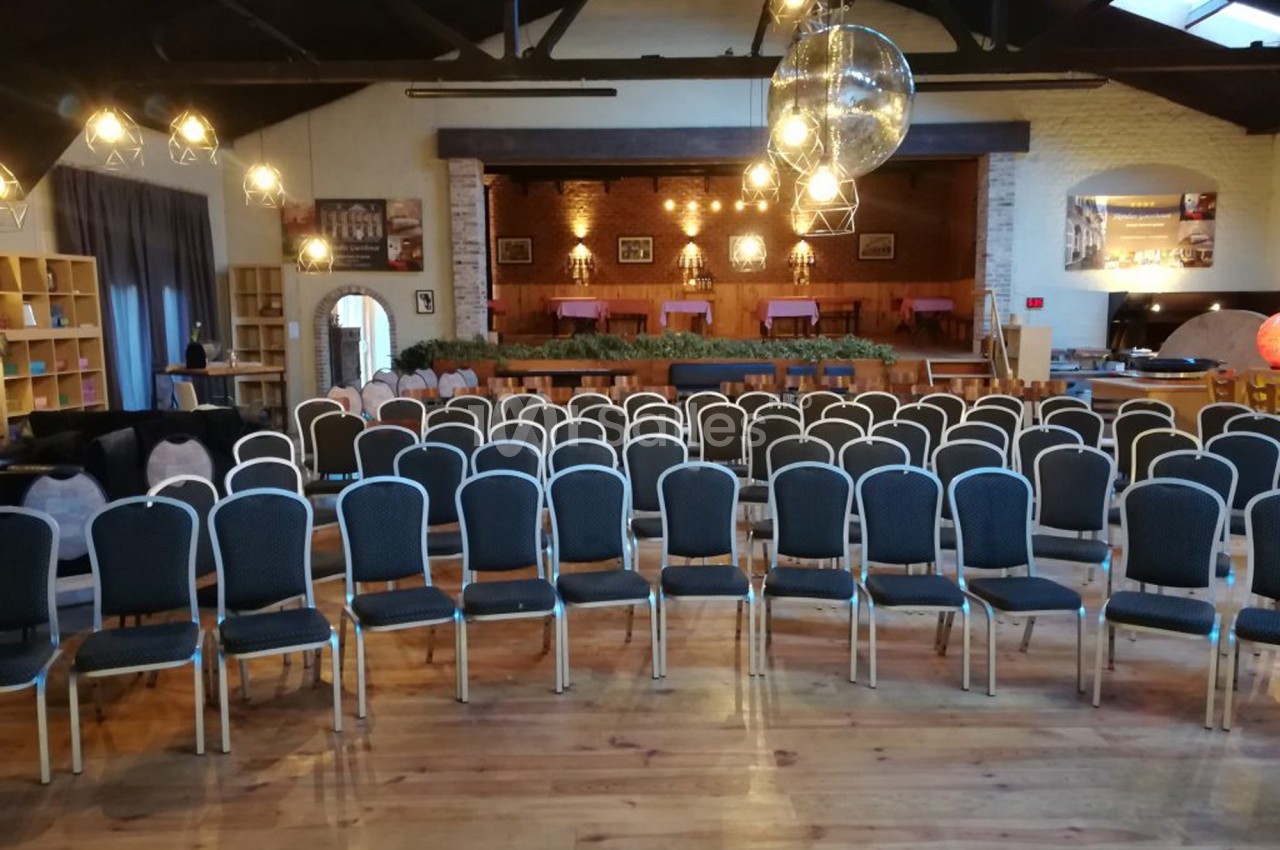 Salle de réception avec rangées de chaises alignées face à une scène décorée de guirlandes lumineuses.