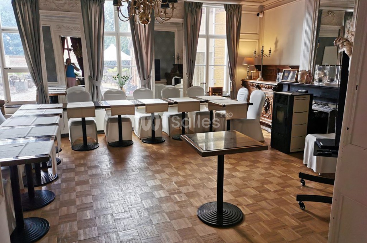 Salle lumineuse avec parquet, tables et chaises disposées en U, décorée de moulures, rideaux et un lustre central.