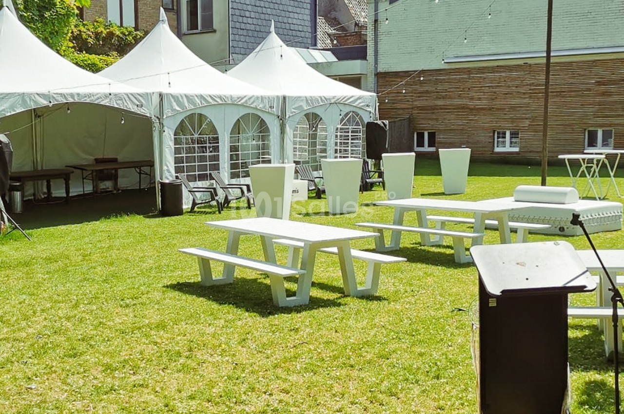 Tables de pique-nique blanches et tentes installées sur une pelouse, entourées de bâtiments en arrière-plan.