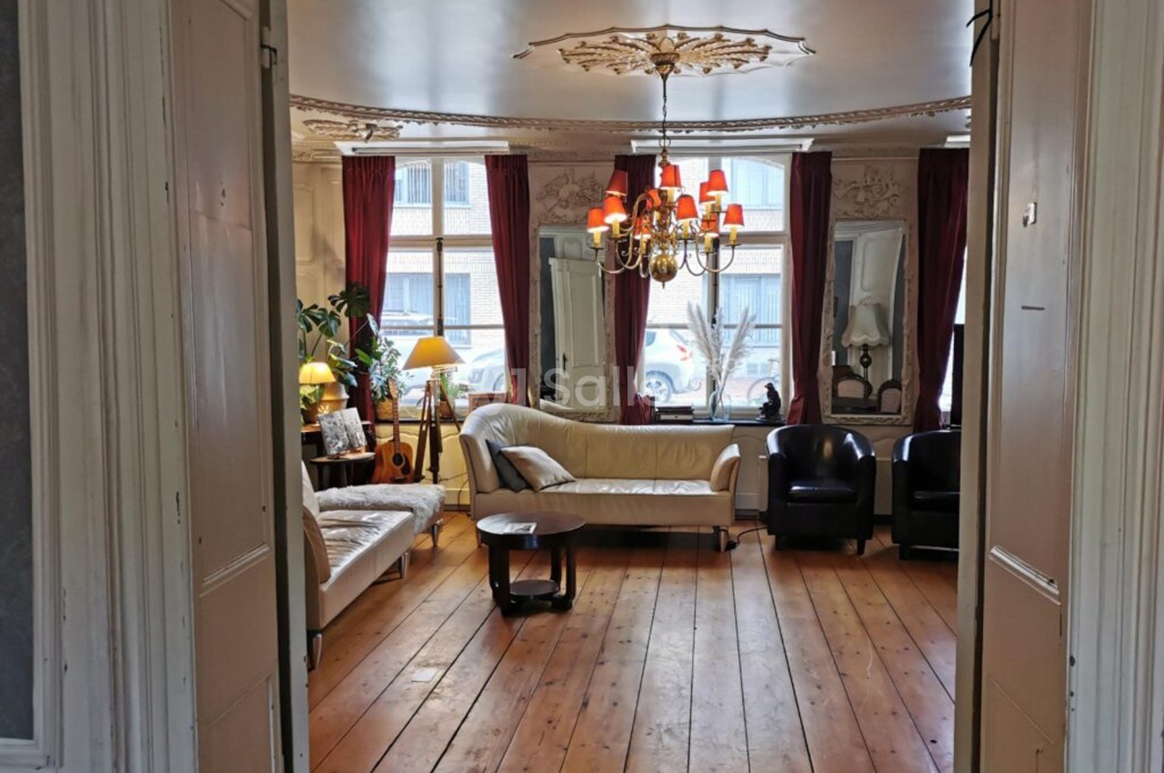 Salon lumineux avec parquet en bois, canapés clairs, fauteuils noirs, lustre central et grandes fenêtres avec rideaux rouges.