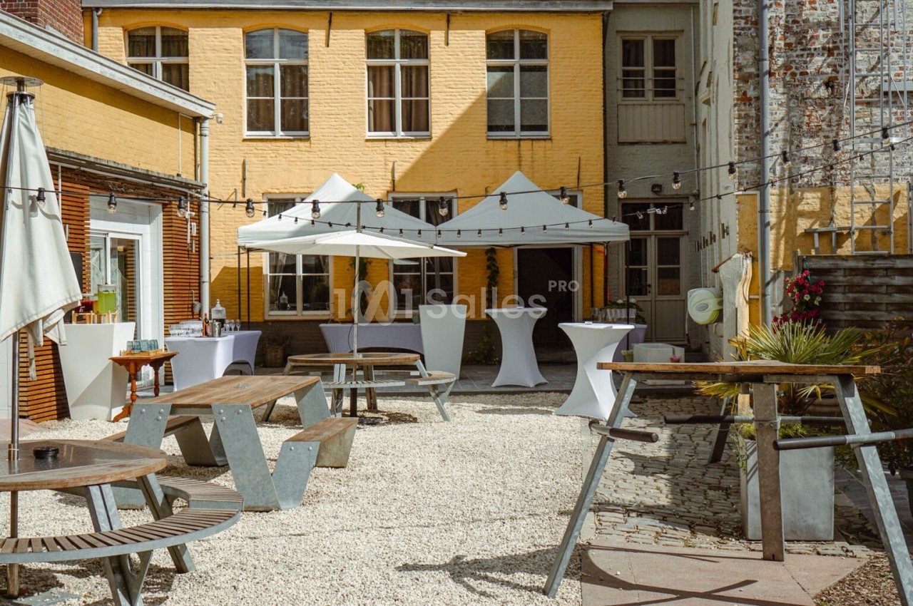 Cour intérieure avec tables en bois, parasols blancs et guirlandes lumineuses, entourée de bâtiments en briques jaunes.