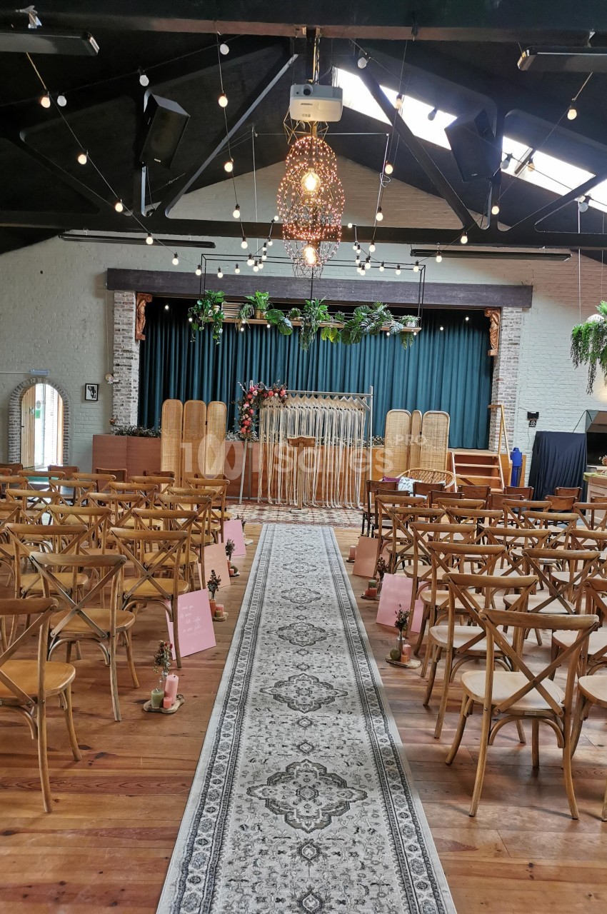 Salle de réception décorée pour une cérémonie, avec des chaises en bois alignées et une arche florale devant une scène.