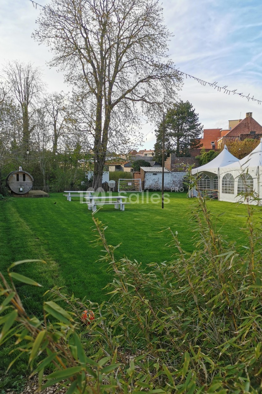 Jardin avec pelouse, tables blanches, tente blanche, tonneau en bois et arbres en arrière-plan sous un ciel dégagé.