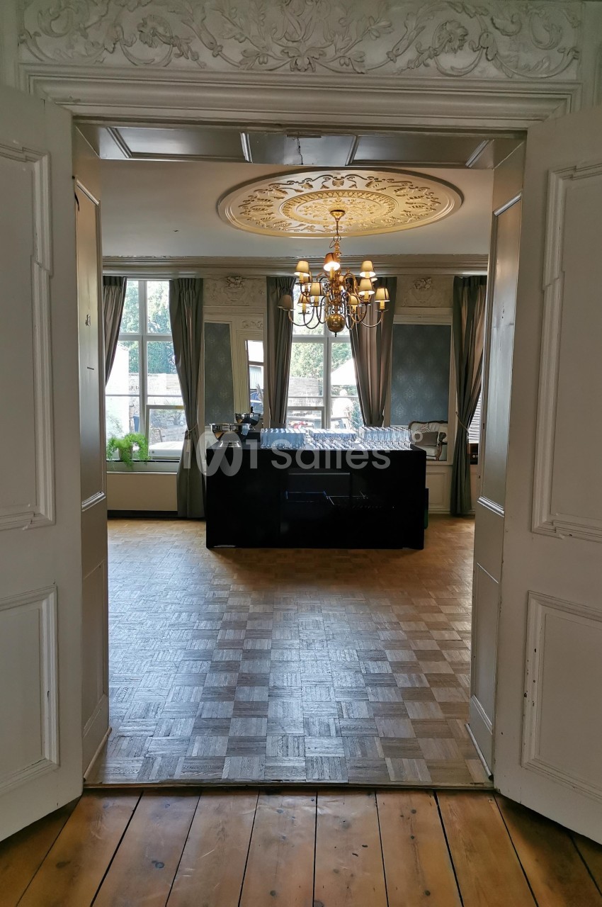 Vue d'une salle élégante avec parquet, lustre, moulures au plafond et grandes fenêtres encadrées de rideaux.