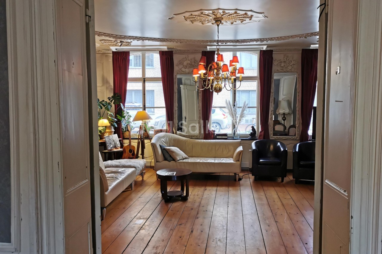 Salon lumineux avec parquet en bois, canapés, fauteuils, lustre et grandes fenêtres encadrées de rideaux rouges.