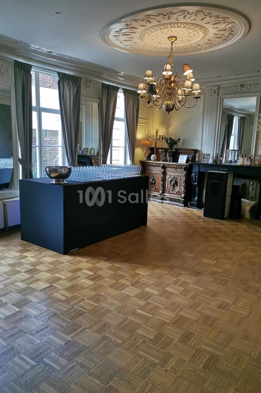 Salle élégante avec parquet, comptoir noir, lustre central et grandes fenêtres encadrées de rideaux.