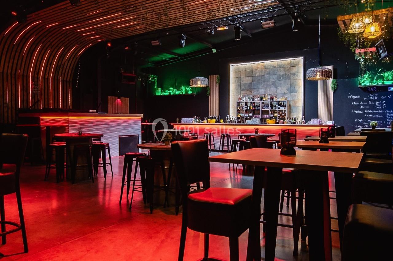 Salle de bar moderne avec éclairage tamisé rouge, tables hautes, chaises noires et comptoir illuminé en arrière-plan.