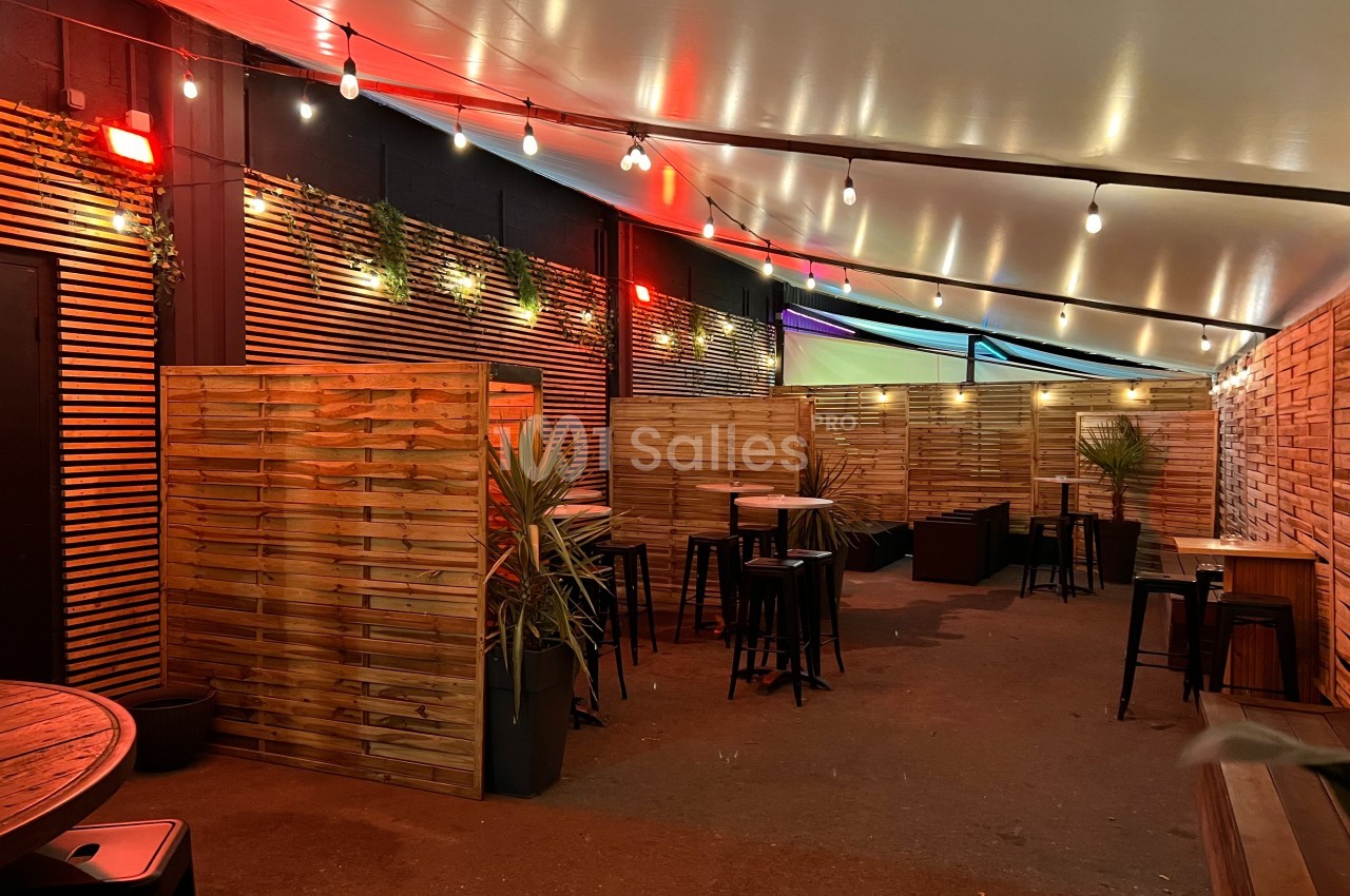 Terrasse extérieure couverte avec éclairage suspendu, séparations en bois, tables hautes et plantes décoratives.