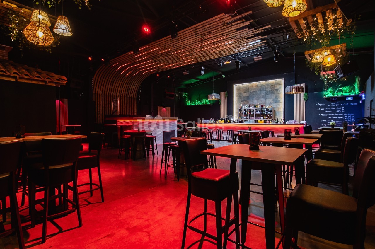 Salle de bar moderne avec éclairage tamisé rouge, tables hautes en bois et comptoir central éclairé.