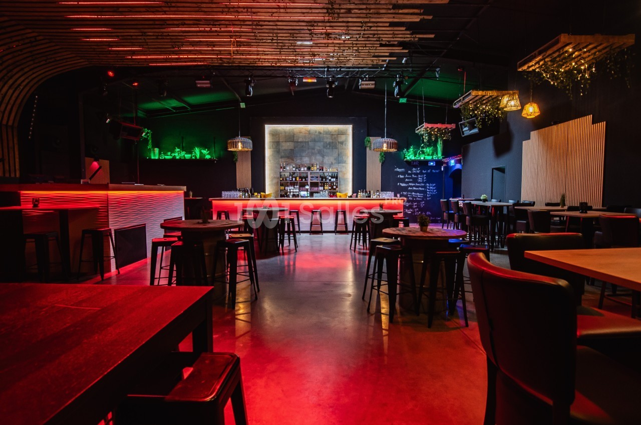 Salle de bar moderne avec éclairage tamisé rouge, comptoir central, tabourets et décoration végétale.