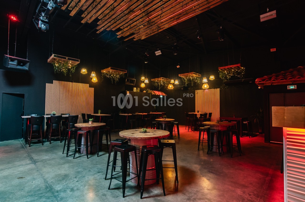 Salle de bar avec tables hautes, tabourets, éclairage tamisé et décoration suspendue végétale.