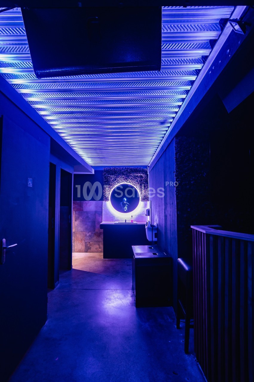Couloir sombre avec éclairage violet, menant à un lavabo éclairé par un miroir rond lumineux.