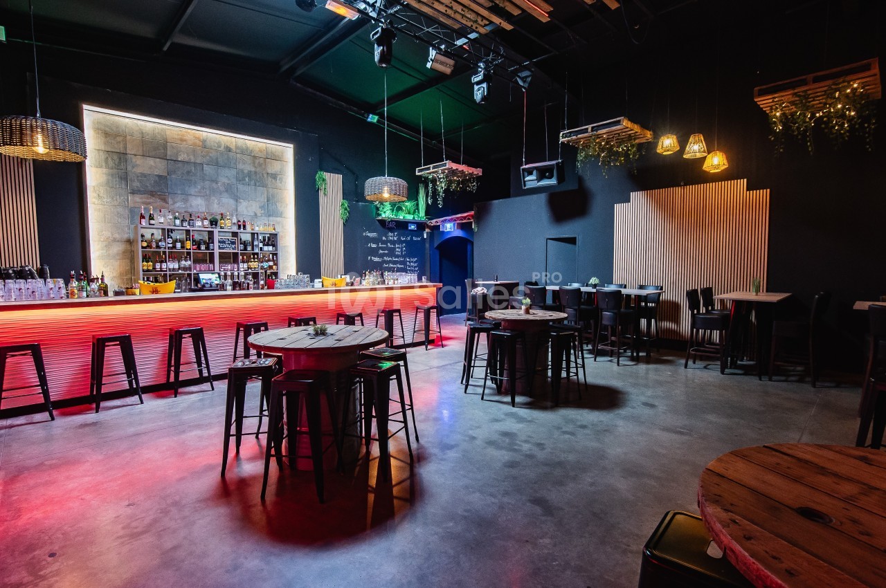 Salle de bar moderne avec comptoir lumineux, tables hautes en bois, chaises noires et éclairage tamisé.