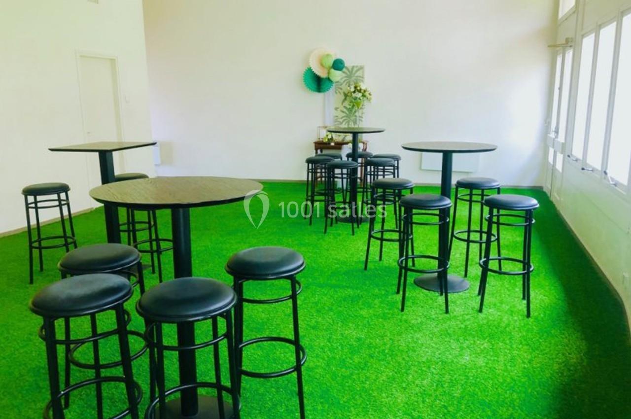 Salle lumineuse avec sol en gazon synthétique, tables hautes noires et tabourets disposés de manière espacée.