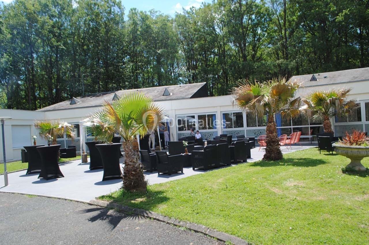 Terrasse aménagée avec tables, chaises et palmiers, devant un bâtiment entouré d'arbres.