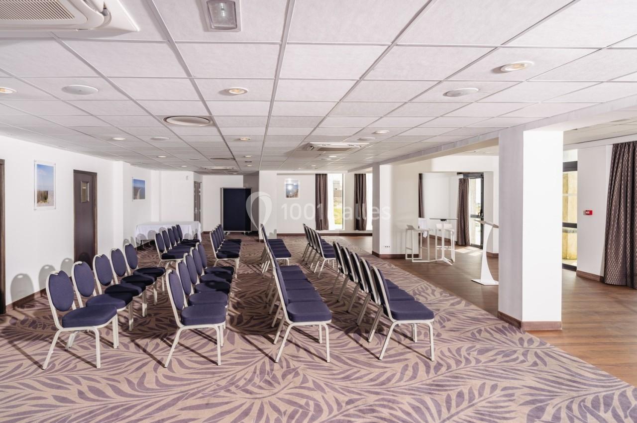 Salle de conférence lumineuse avec des rangées de chaises bleues alignées sur un sol à motif, entourée de fenêtres et de…