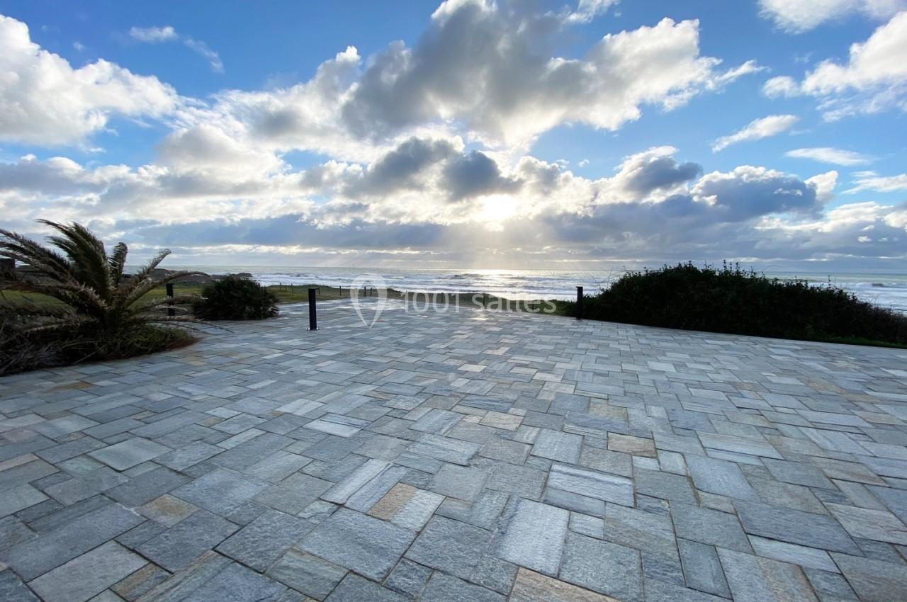 Vue d'une terrasse pavée avec un palmier, des buissons et l'océan en arrière-plan sous un ciel partiellement nuageux.