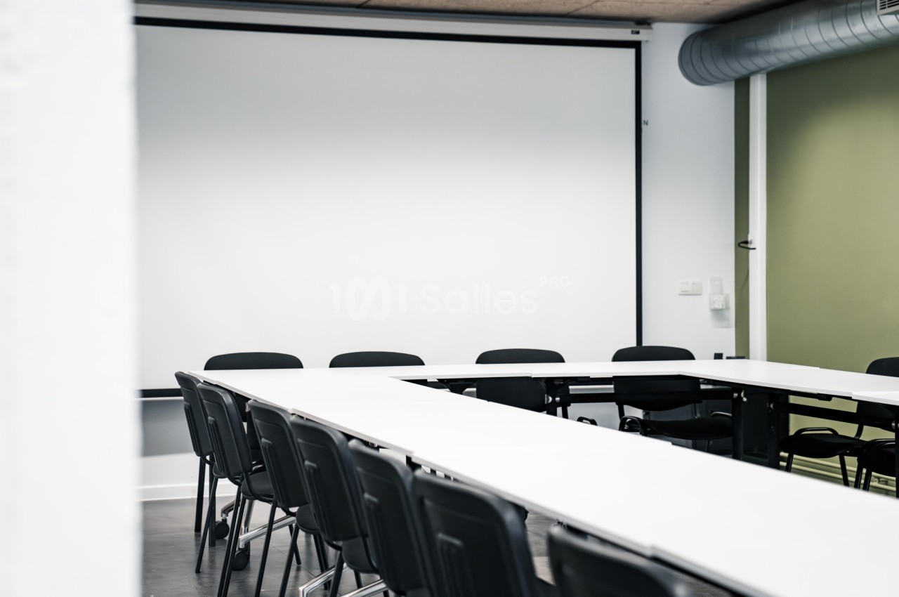 Salle de réunion vide avec des tables blanches disposées en U et des chaises noires, devant un écran de projection.