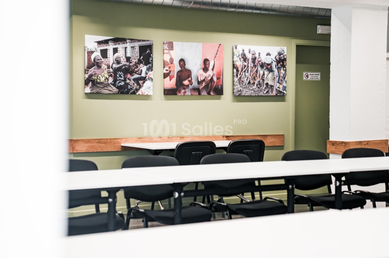 Salle de réunion avec des tables blanches, des chaises noires et des photos accrochées sur un mur vert.