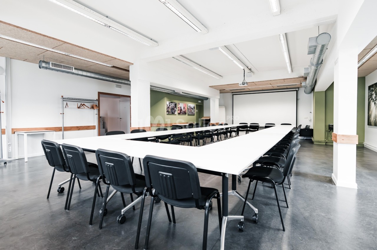 Salle de réunion lumineuse avec tables disposées en U, chaises noires et écran de projection en fond.