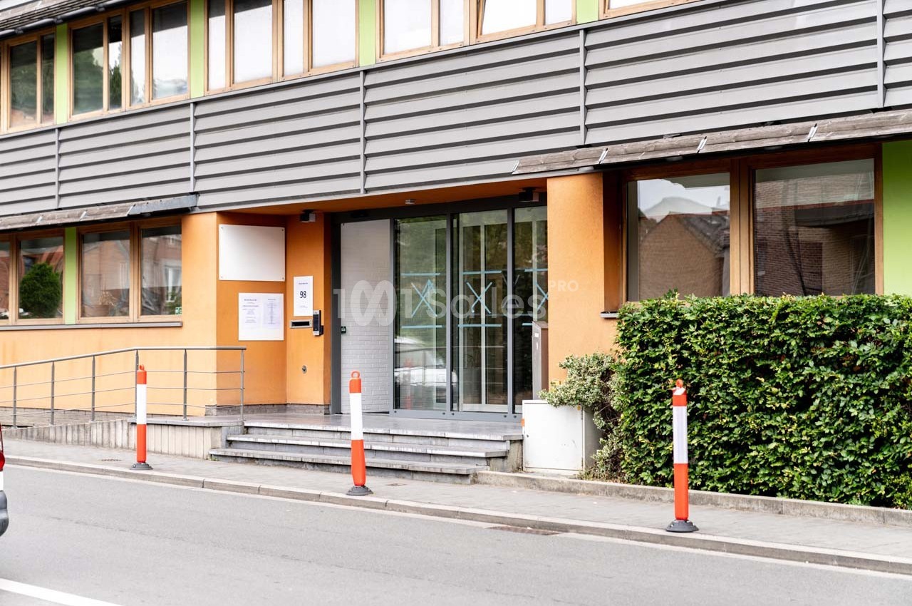 Entrée d'un bâtiment moderne avec rampe d'accès, porte vitrée et poteaux de sécurité sur le trottoir.