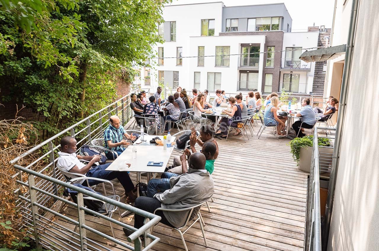Personnes assises à des tables sur une terrasse en bois, entourée de verdure et d'immeubles modernes en arrière-plan.
