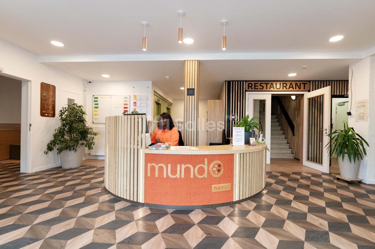 Accueil avec comptoir en bois, employée assise, plantes décoratives et signalétique vers un restaurant en arrière-plan.
