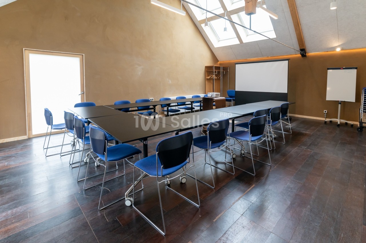 Salle de réunion lumineuse avec tables disposées en U, chaises bleues, tableau blanc et écran de projection.