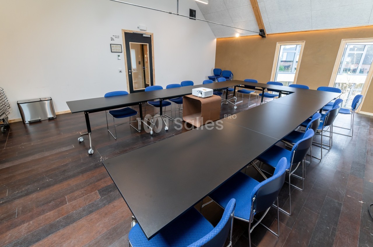 Salle de réunion lumineuse avec tables noires en U, chaises bleues et projecteur sur un meuble central.