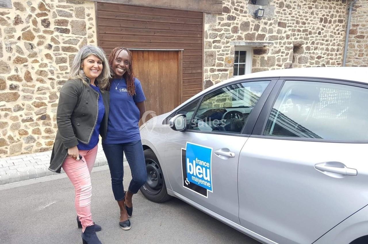 Deux femmes souriantes posent devant une voiture grise marquée ’France Bleu’, stationnée devant un bâtiment en pierre.