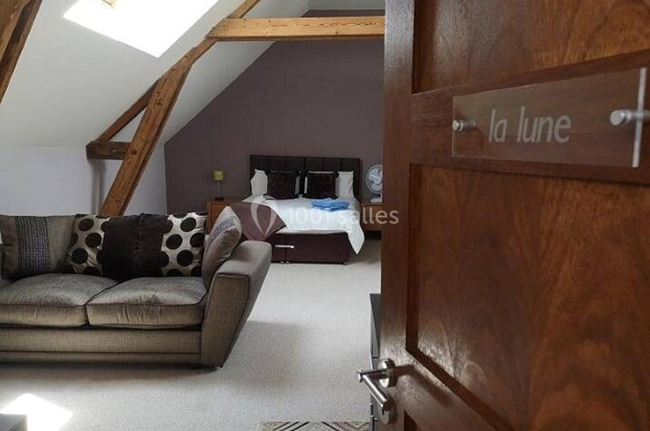Vue d'une chambre mansardée avec un lit double, un canapé, des poutres apparentes et une porte en bois ouverte.